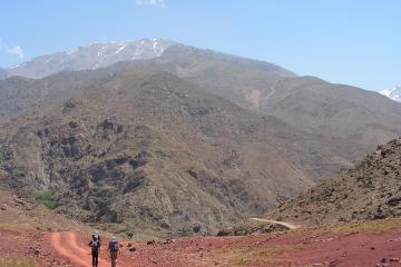 Two hikers with backpacks walk along a reddish dirt road in a mountainous landscape, with a large, snow-capped peak visible in the background under a clear blue sky.