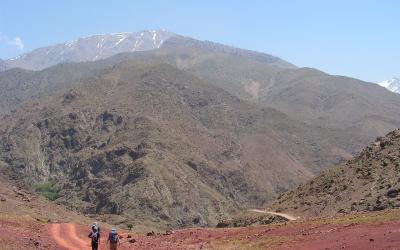 Two hikers with backpacks walk along a reddish dirt road in a mountainous landscape, with a large, snow-capped peak visible in the background under a clear blue sky.