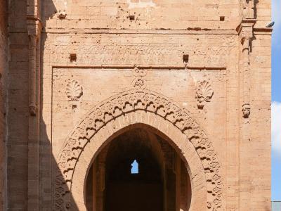 A large, ornate sandstone gateway with a horseshoe arch and crenellated parapet, featuring visitors entering through the archway under a blue sky.