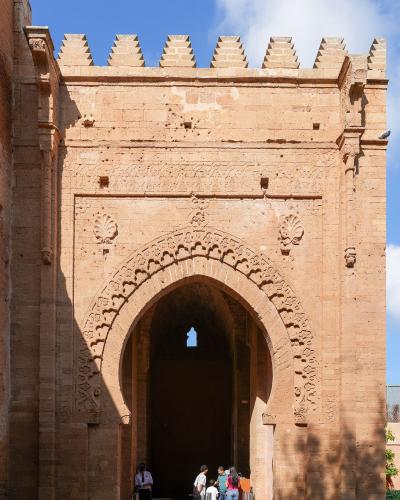 A large, ornate sandstone gateway with a horseshoe arch and crenellated parapet, featuring visitors entering through the archway under a blue sky.