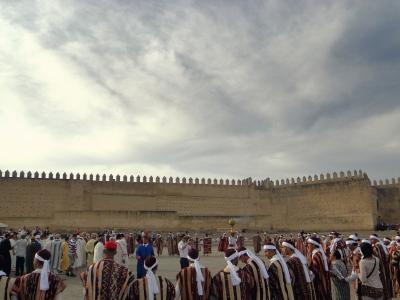 A large group of people in traditional striped robes and white head coverings are gathered in a paved square in front of a massive, crenellated earthen wall under a cloudy sky.