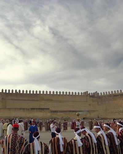 A large group of people in traditional striped robes and white head coverings are gathered in a paved square in front of a massive, crenellated earthen wall under a cloudy sky.