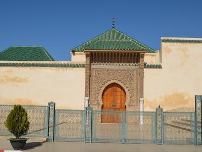 A beige stone wall with a large, ornately carved horseshoe archway and wooden doors, topped by a green-tiled roof, with a metal fence in the foreground.