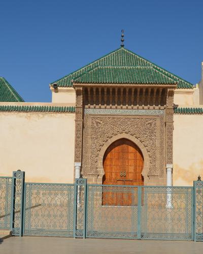 A beige stone wall with a large, ornately carved horseshoe archway and wooden doors, topped by a green-tiled roof, with a metal fence in the foreground.