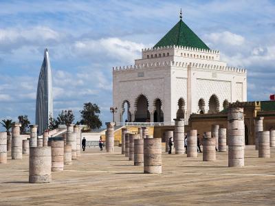 A low-angle view of a large white mausoleum with a green pyramidal roof and Moorish arches, situated in a plaza filled with ancient stone columns, with a modern skyscraper visible in the background.