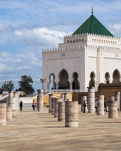 A low-angle view of a large white mausoleum with a green pyramidal roof and Moorish arches, situated in a plaza filled with ancient stone columns, with a modern skyscraper visible in the background.