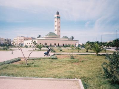 A wide-angle photograph of a large mosque with a tall, square minaret and a green-tiled roof, situated in a landscaped park area with palm trees and benches under a partly cloudy sky.