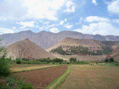 A landscape view of agricultural fields in the foreground leading to arid, rolling hills and a large mountain range in the background, with a small structure visible on a hilltop.