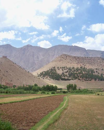 A landscape view of agricultural fields in the foreground leading to arid, rolling hills and a large mountain range in the background, with a small structure visible on a hilltop.