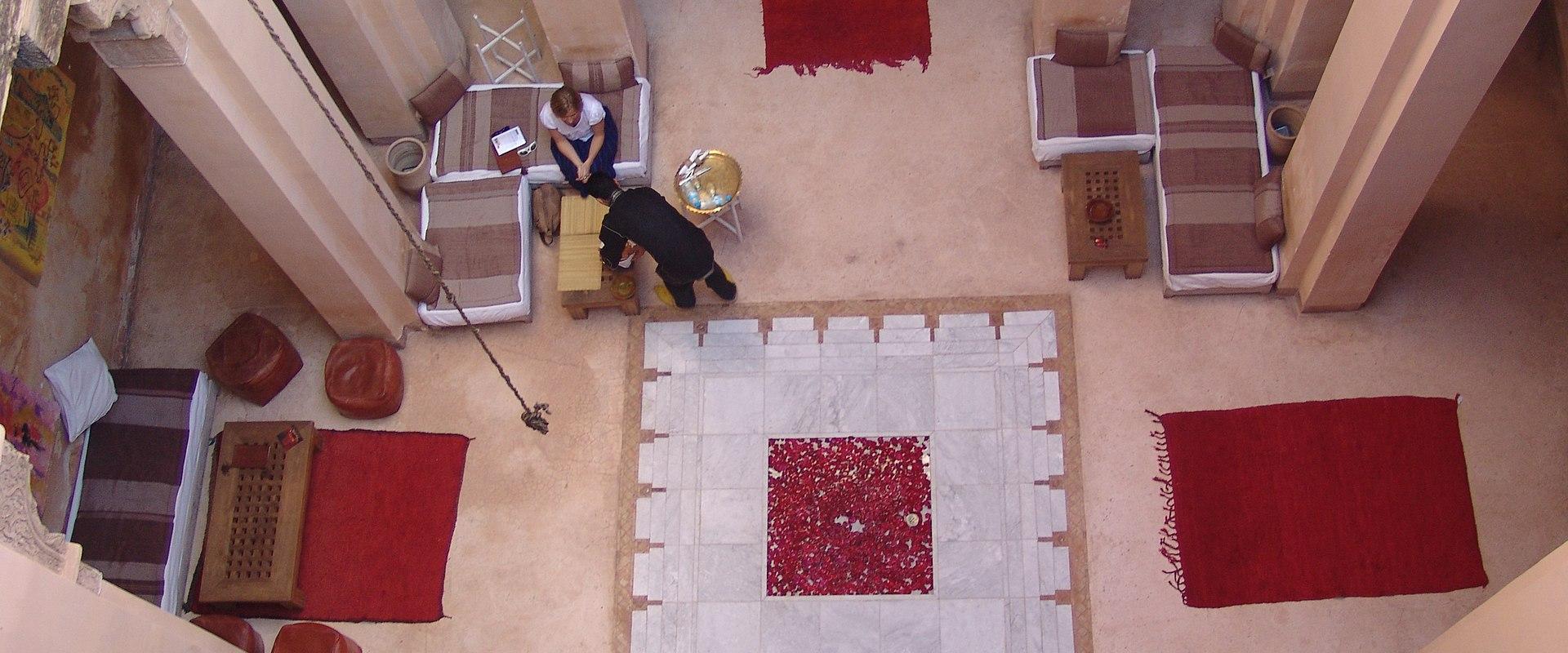 A high-angle view of a traditional Moroccan riad courtyard with white marble flooring, red rugs, cushioned seating, and people relaxing.