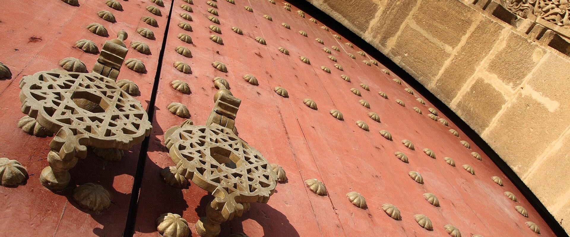 A close-up view of a massive, red-painted wooden door studded with metal bosses and featuring large, ornate iron knockers, set within a stone archway with carved stonework above.