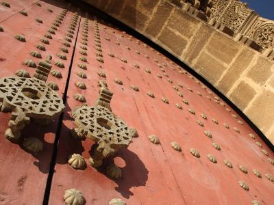 A close-up view of a massive, red-painted wooden door studded with metal bosses and featuring large, ornate iron knockers, set within a stone archway with carved stonework above.