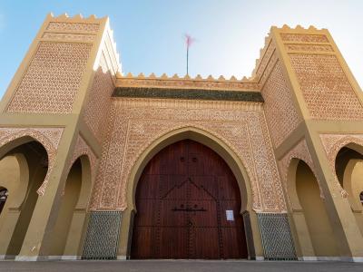 A low-angle shot of a large, ornate entrance gate with two flanking towers, intricate geometric tilework (zellige), and a large wooden door topped by a Moroccan flag.