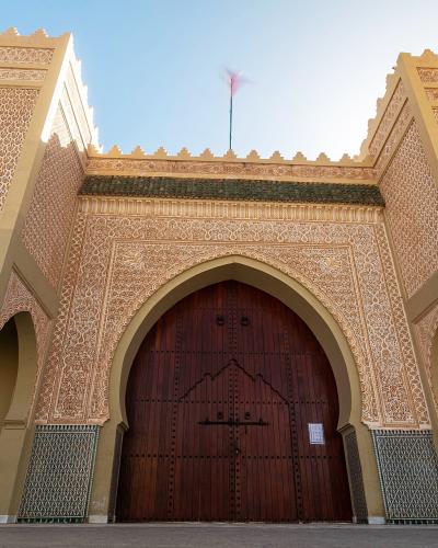 A low-angle shot of a large, ornate entrance gate with two flanking towers, intricate geometric tilework (zellige), and a large wooden door topped by a Moroccan flag.
