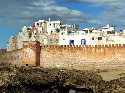 A view of the fortified sea walls of a coastal old town with white buildings behind them and people sorting fishing nets on the rocky shore in the foreground.