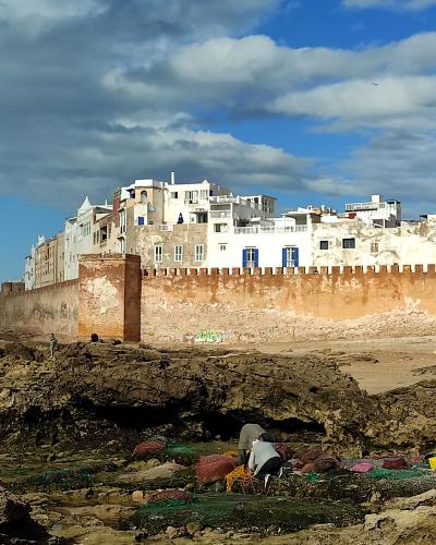 A view of the fortified sea walls of a coastal old town with white buildings behind them and people sorting fishing nets on the rocky shore in the foreground.