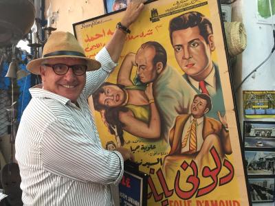 A man in a hat and striped shirt stands indoors in a shop filled with antiques and vintage items, holding a large framed vintage Arabic/French movie poster for "Folie d'Amour" which lists "CASABLANCA" at the bottom.