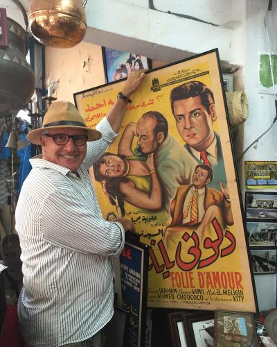 A man in a hat and striped shirt stands indoors in a shop filled with antiques and vintage items, holding a large framed vintage Arabic/French movie poster for "Folie d'Amour" which lists "CASABLANCA" at the bottom.