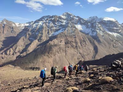 A group of hikers with backpacks walking on a rocky, high-altitude trail with a large, snow-dusted mountain peak in the background under a blue sky.