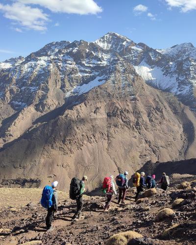 A group of hikers with backpacks walking on a rocky, high-altitude trail with a large, snow-dusted mountain peak in the background under a blue sky.