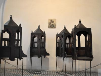 Four ornate, dark wooden palanquins (sedan chairs) displayed on metal stands inside a museum room with white walls and a checkered tile floor.