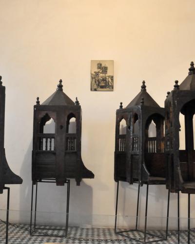 Four ornate, dark wooden palanquins (sedan chairs) displayed on metal stands inside a museum room with white walls and a checkered tile floor.
