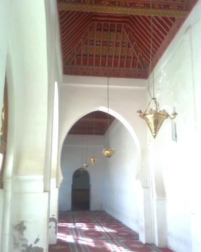 An interior view of a prayer hall featuring white walls, horseshoe arches, a red patterned carpet, and an ornate, painted wooden ceiling with hanging brass lanterns.