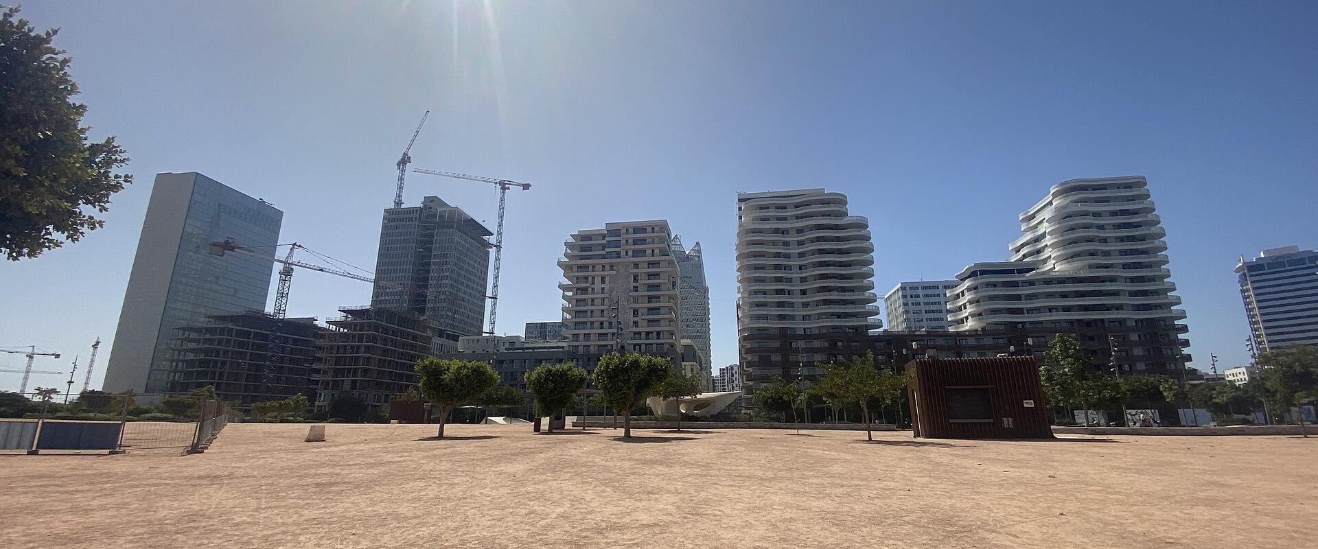 A wide, sandy open area in the foreground with several modern high-rise buildings and construction cranes in the background under a bright sun.