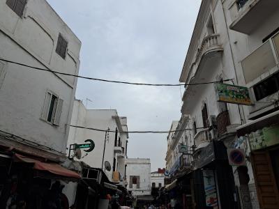A narrow street scene in a densely built urban area with whitewashed buildings, overhead electrical wires, and various shop signs including Arabic script and a pharmacy symbol.