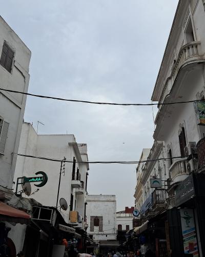 A narrow street scene in a densely built urban area with whitewashed buildings, overhead electrical wires, and various shop signs including Arabic script and a pharmacy symbol.