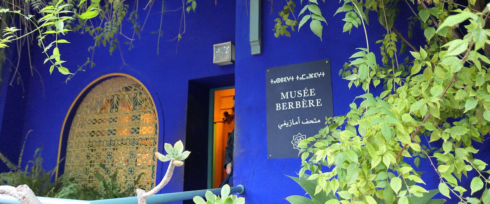 A blue-walled building entrance with a sign reading "MUSÉE BERBÈRE" and Arabic text, surrounded by green foliage and succulents.