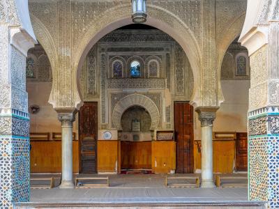 The interior of a prayer hall featuring horseshoe arches, intricate stucco carvings, marble columns, and a mihrab niche, with wooden paneling and prayer mats on the floor.