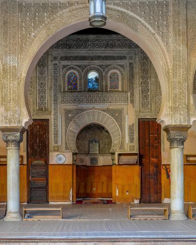 The interior of a prayer hall featuring horseshoe arches, intricate stucco carvings, marble columns, and a mihrab niche, with wooden paneling and prayer mats on the floor.