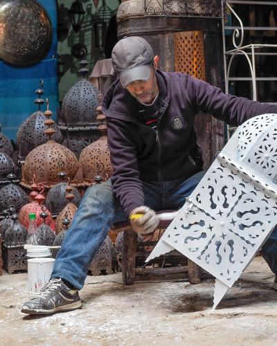 A man wearing a cap and work clothes sits on a low stool in an outdoor workshop or shop front, painting a large, white, ornate metal lantern with a brush, surrounded by numerous other finished and unfinished metal lanterns and lamps.