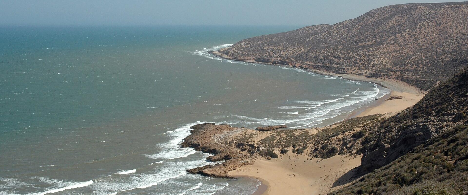 A high-angle view of a long, sandy beach curving along a coastline with arid, scrub-covered hills and rocky outcrops under a clear sky.