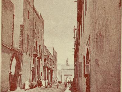 A sepia-toned photograph of a narrow street in a walled city, featuring buildings with arched doorways and balconies, with figures in traditional dress and a church tower visible in the distance.