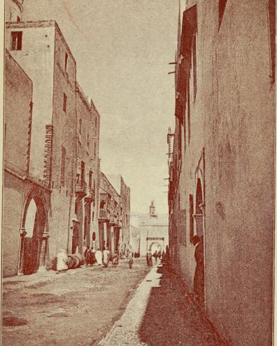 A sepia-toned photograph of a narrow street in a walled city, featuring buildings with arched doorways and balconies, with figures in traditional dress and a church tower visible in the distance.