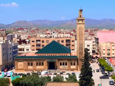 A large mosque with a tall, square minaret and a distinctive green pyramidal roof, situated in a dense urban area with mountains in the background.