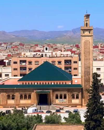 A large mosque with a tall, square minaret and a distinctive green pyramidal roof, situated in a dense urban area with mountains in the background.