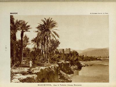 A vintage sepia photograph showing a riverbank with palm trees and a traditional fortified village (ksar) in the background, with printed text identifying the location as "KSAR-ES-SOUK, dans le Tafilalet (Sahara Marocain)".