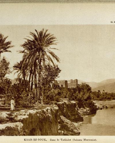 A vintage sepia photograph showing a riverbank with palm trees and a traditional fortified village (ksar) in the background, with printed text identifying the location as "KSAR-ES-SOUK, dans le Tafilalet (Sahara Marocain)".