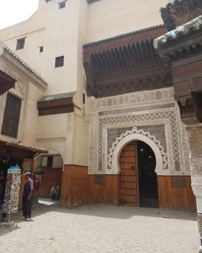 The image shows the ornate, arched entrance facade of a historic building with intricate zellige tilework and carved wood, situated in a paved courtyard with a souvenir stand to the left.