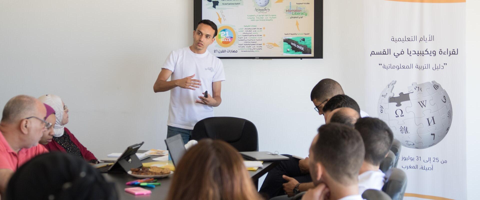 A man in a white t-shirt stands presenting to a seated group in a classroom or meeting room, with a large screen displaying a map and a vertical banner featuring the Wikipedia logo and Arabic text.