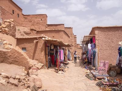 A high-angle view of a fortified earthen village (ksar) with distinctive tower-like structures, situated next to a dry riverbed and surrounded by palm groves and arid hills.