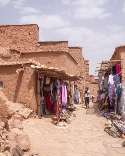 A high-angle view of a fortified earthen village (ksar) with distinctive tower-like structures, situated next to a dry riverbed and surrounded by palm groves and arid hills.