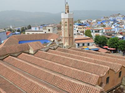 A high-angle view of a large mosque with a terracotta-tiled roof and a square minaret, set against the backdrop of the blue-painted buildings of Chefchaouen, Morocco.