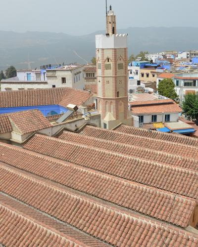A high-angle view of a large mosque with a terracotta-tiled roof and a square minaret, set against the backdrop of the blue-painted buildings of Chefchaouen, Morocco.