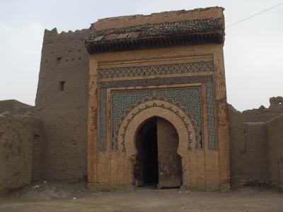An ornate, arched gateway (bab) featuring intricate zellige tilework and carved stucco, set within a larger earthen wall structure.