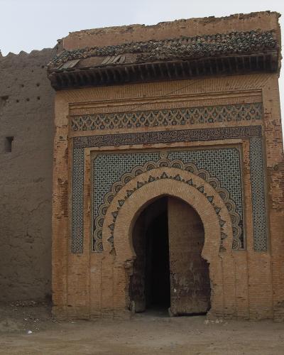 An ornate, arched gateway (bab) featuring intricate zellige tilework and carved stucco, set within a larger earthen wall structure.