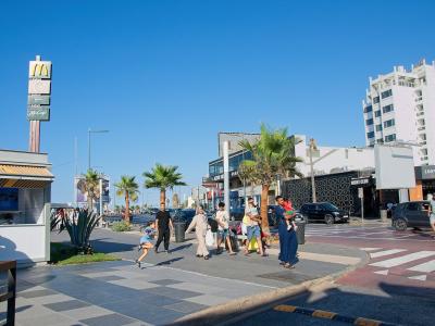 A sunny outdoor street scene showing a paved promenade with pedestrians, palm trees, a McDonald's sign, and modern buildings along a coastal road.